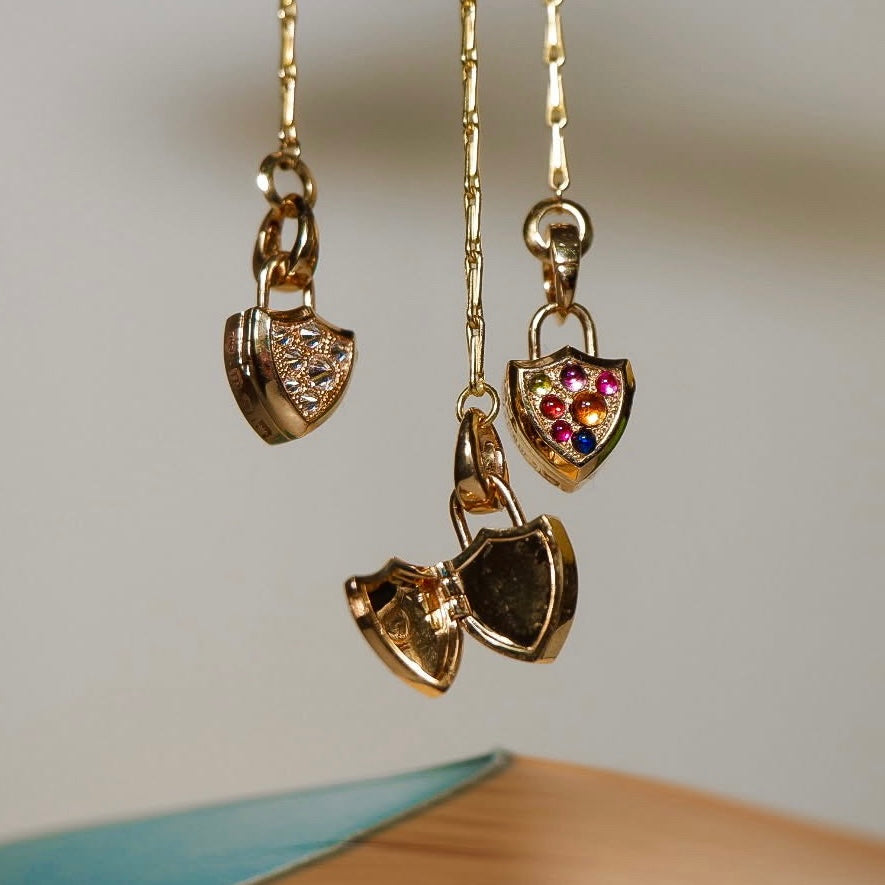 Three gold shield-shaped pendant necklaces hang against a soft beige background. Two pendants are closed , one shows colorful cabochon gems and one shows pavé diamonds on the front, and one is open,