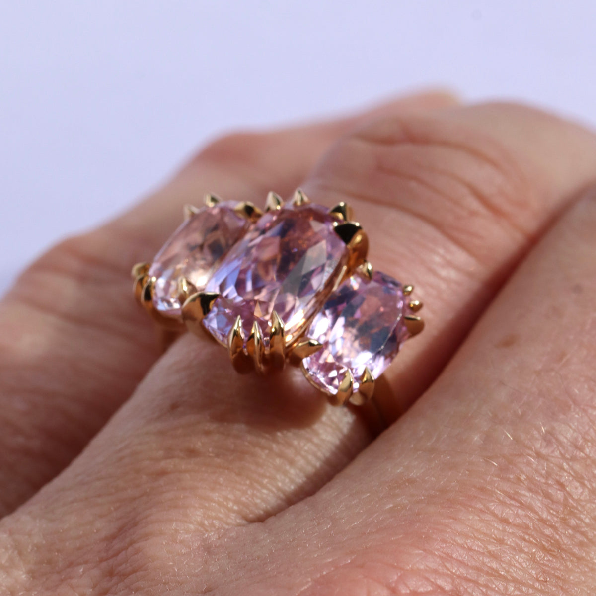 close up of models hand wearing a large cocktail ring with three pink Kunzite gemstones and asymmetric knife edged gold setting, by Tamsin. Rasor, against a white background