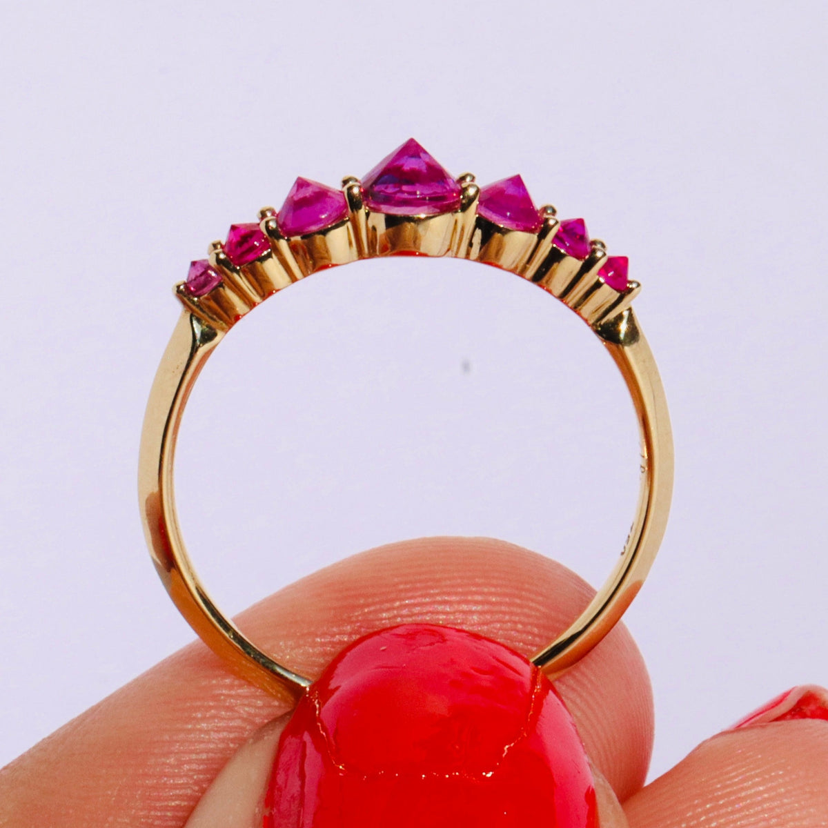 Spiky seven stone ruby and gold ring , held upright between models thumb and forefinger against white background by Tamsin Rasor
