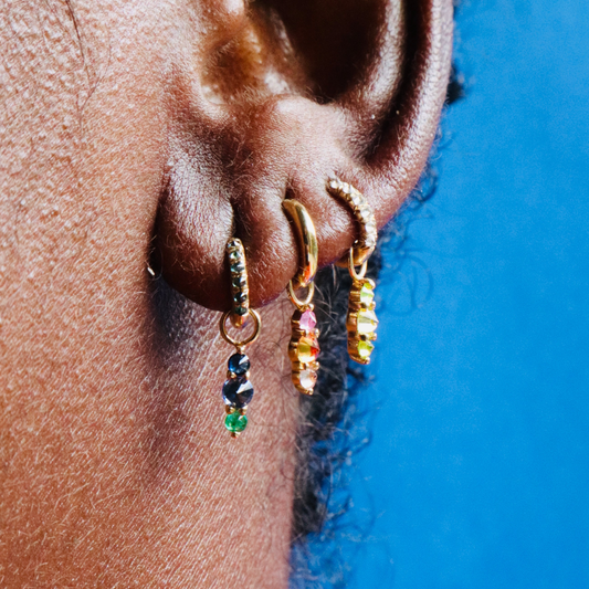 Three Mini Barbed Diamond Chunky huggie hoop earrings with colorful gemstone charms on models ear against blue background