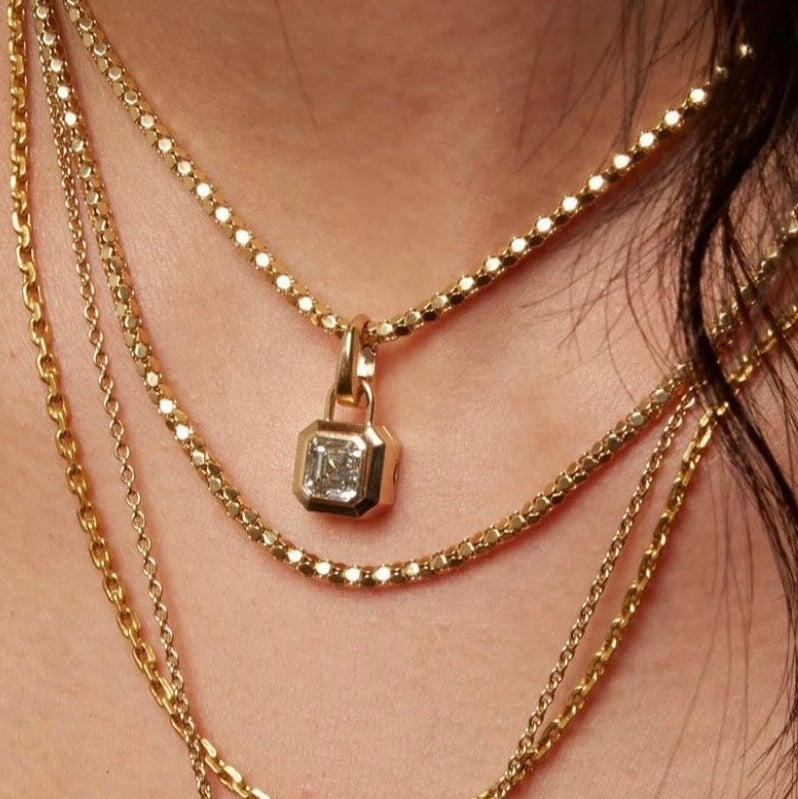 Gold square shaped padlock inspired locket pendant with Asscher cut white diamond in the center, on textured gold chain layered with three other gold chains, on model, By Tamsin Rasor
