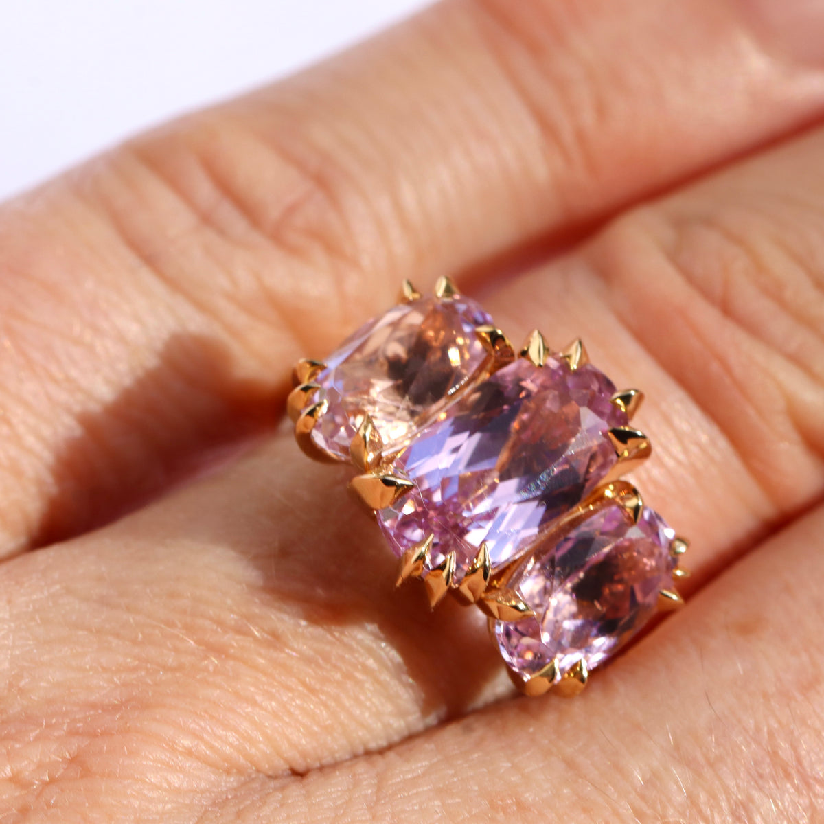 close up of models hand wearing a large cocktail ring with three pink Kunzite gemstones and asymmetric knife edged gold setting, by Tamsin. Rasor, against a white background