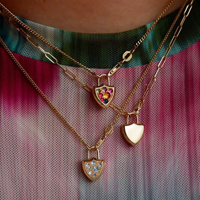 Close up of a person wearing thren gold chain necklaces with three shield shaped charms, one solid gold, one with colorful gemstone accents, one with diamond accents, model wears colorful tie-dye top.