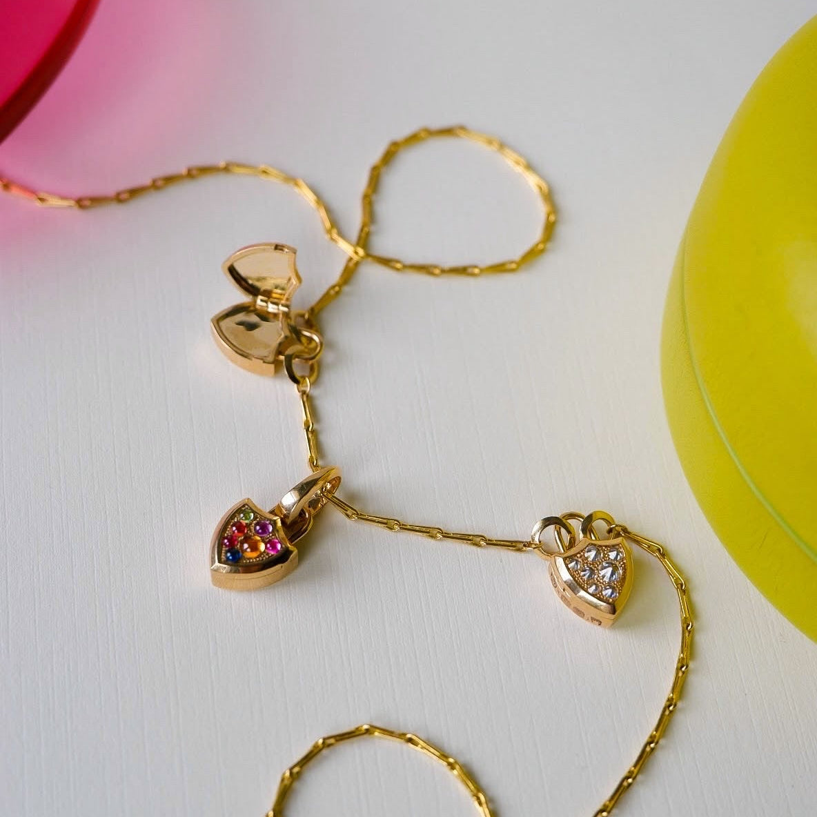 A gold chain necklace with three shield shaped charms, one with colorful gemstone accents, one with diamond accents, displayed on a white surface with decorative colorful props.