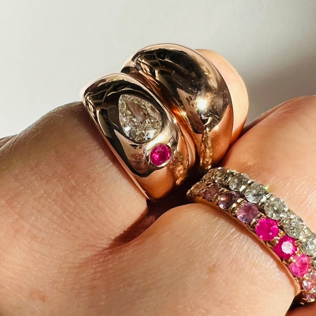 Close up image of a hand on white back ground showing two domed rose gold rings on pinky finger. One has a pear shaped diamond and pink sapphire set in it the other is solid gold. White diamond eternity ring and ombré pink sapphire eternity ring stacked on ring finger.