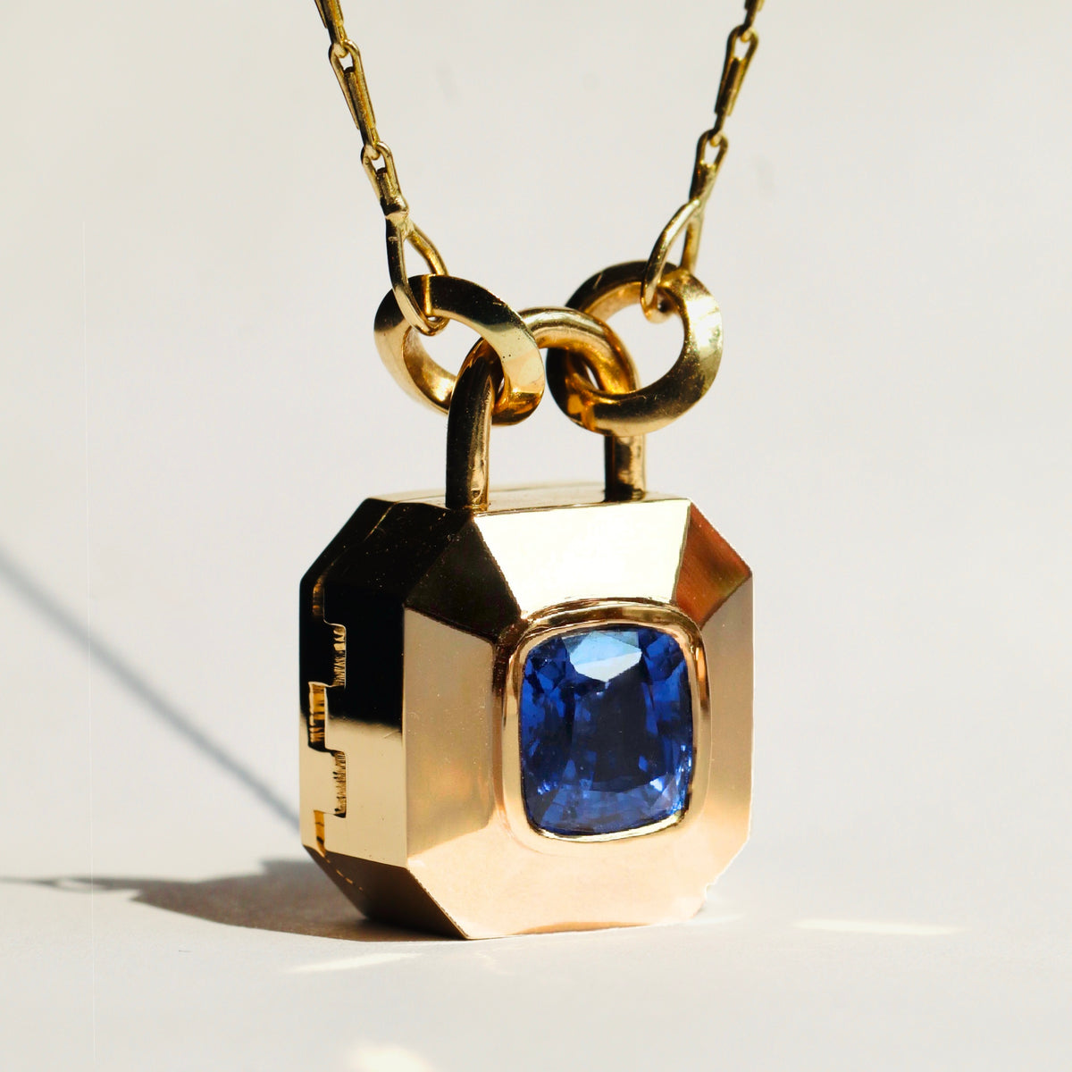 A gold padlock-shaped locket pendant with a blue cushion cut sapphire, on gold chain set against a plain white background, by Tamsin Rasor Fine Jewelry