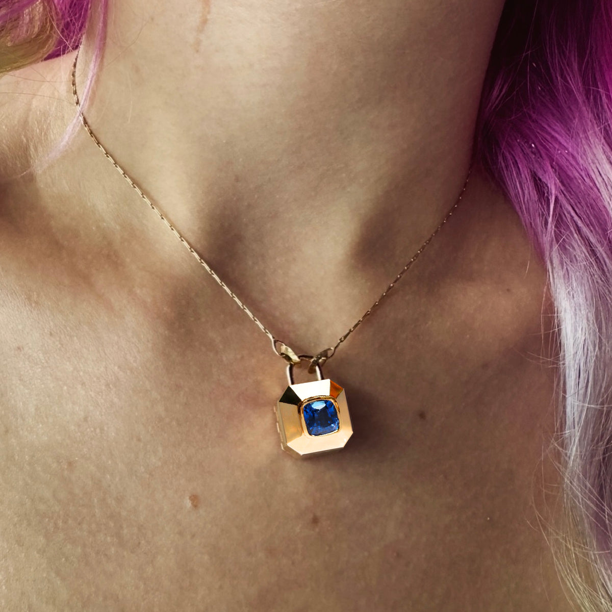 A gold padlock-shaped locket pendant on gold chain with a blue cushion cut sapphire, shown on model with pink hair, by Tamsin Rasor Fine Jewelry