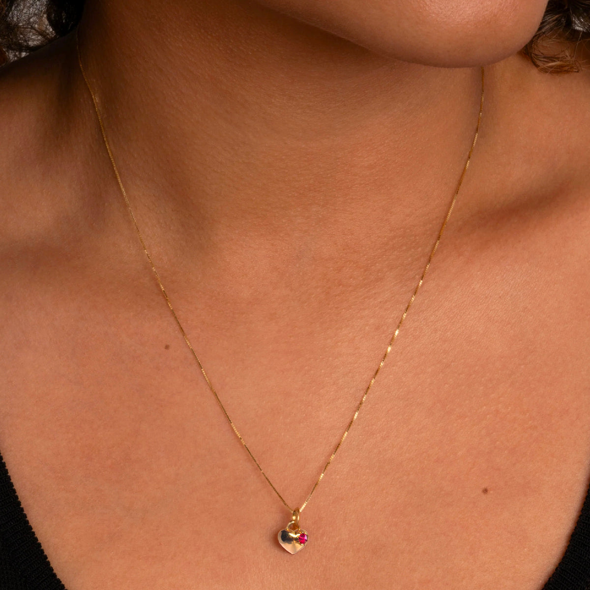 Gold heart-shaped pendant on a chain, with a single star set spiked ruby gemstone on the right hand side. Shown around models neck
