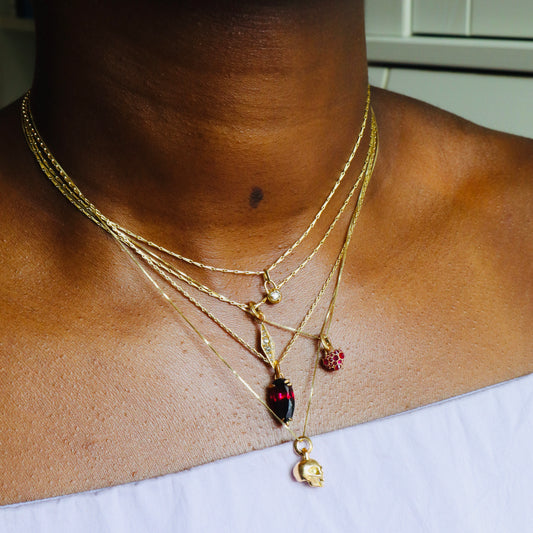 Five stacked gold necklaces on model including mini diamond compass, barbed diamond obelisk, barbed ruby heart, garnet pear drop, mini diamond skull