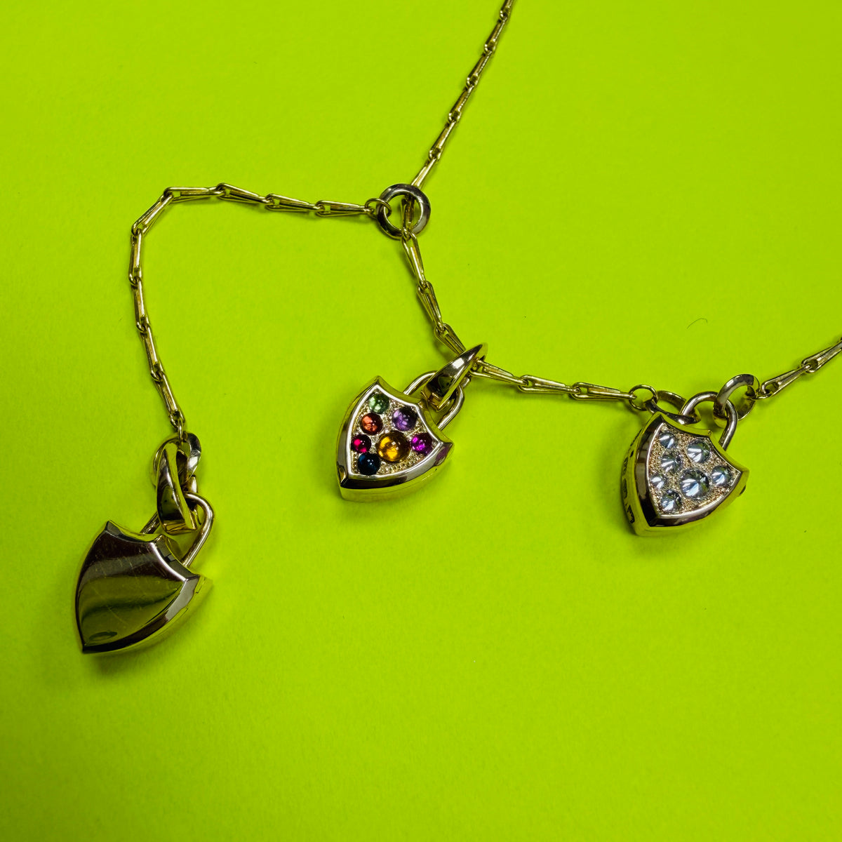 Gold chain necklace featuring three shield-shaped locket charms: one plain polished gold, one with small multicolor cabochons, and one with linverted diamond pavé, displayed on a bright lime green background. By Tamsin Rasor