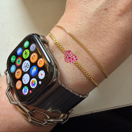 Gold bracelet with a heart-shaped ruby charm by Tamsin Rasor shown on models wrist stacked with gold chain bracelets and a smart watch