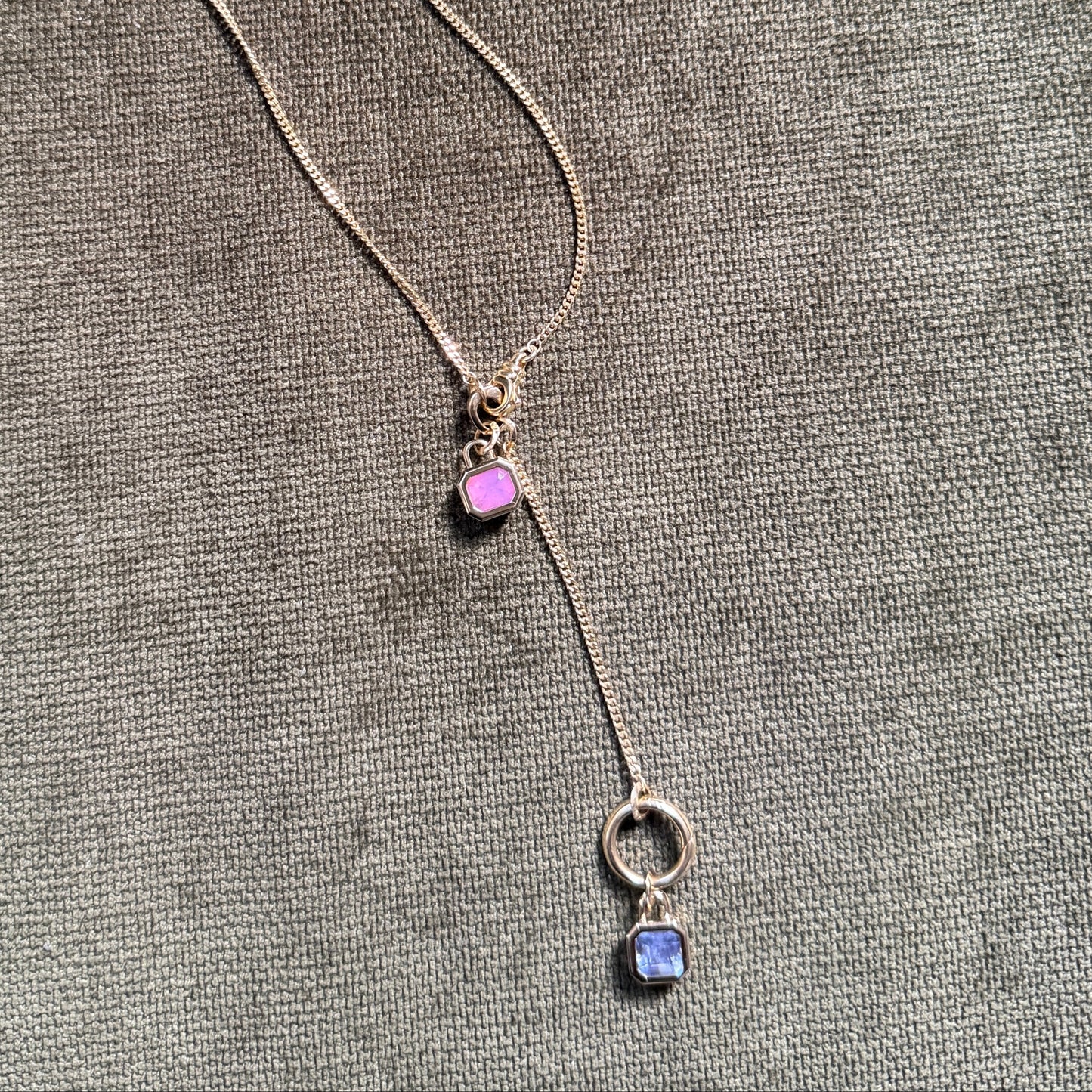 bezel set gemstone pendants on gold chain with charm clips. One pin Spinel and one lavender sapphire. Against olive green velvet background