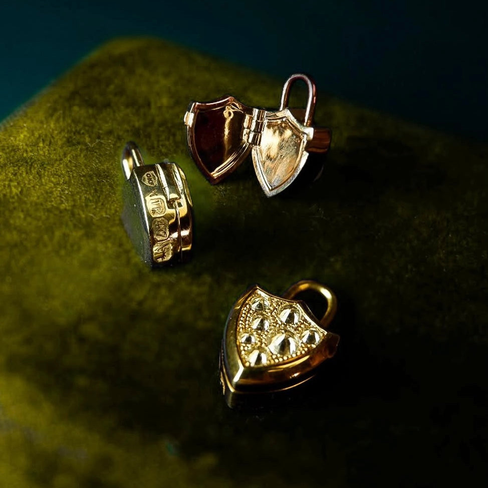 Three small shield-shaped gold pendants , one with spiked diamond pavé, by Tamsin Rasor displayed on a dark green velvet surface against a deep blue background.