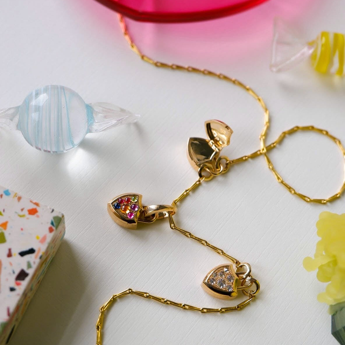 A gold chain necklace with three shield shaped charms, one with colorful gemstone accents, one with diamond accents, displayed on a white surface with decorative colorful props.