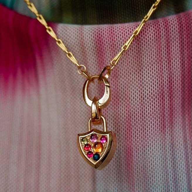 A close-up of a gold necklace worn on a person's neck, featuring a clip bail holding a small shield shaped locket pendant with rainbow cabochon pavé, over a teal & pink tie-dye mesh shirt.