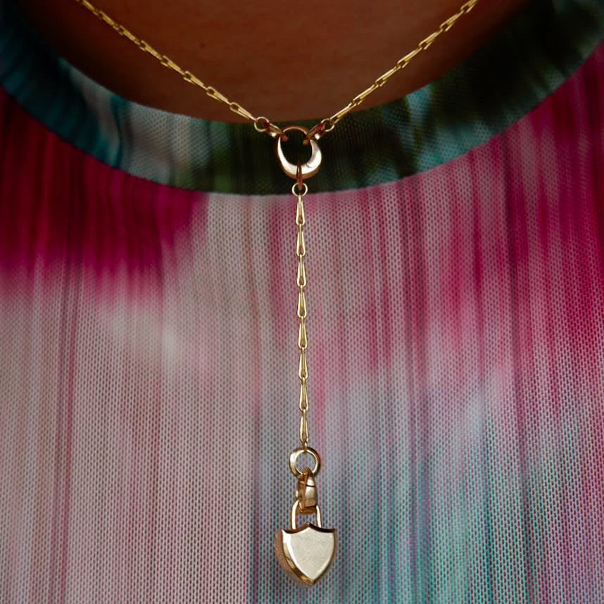 A close-up of a gold necklace worn on a person's neck, featuring a Y-shaped chain design with a mini shield shaped locket pendant, made of solid gold,  over a teal & pink tie-dye mesh shirt.