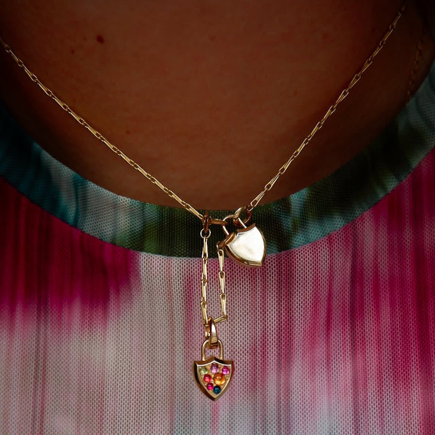 A close-up of a gold necklace worn on a person's neck, featuring a looped chain design with two small shield shaped locket pendants, one solid gold and one gold with rainbow cabochon pavé, over a teal & pink tie-dye mesh shirt.