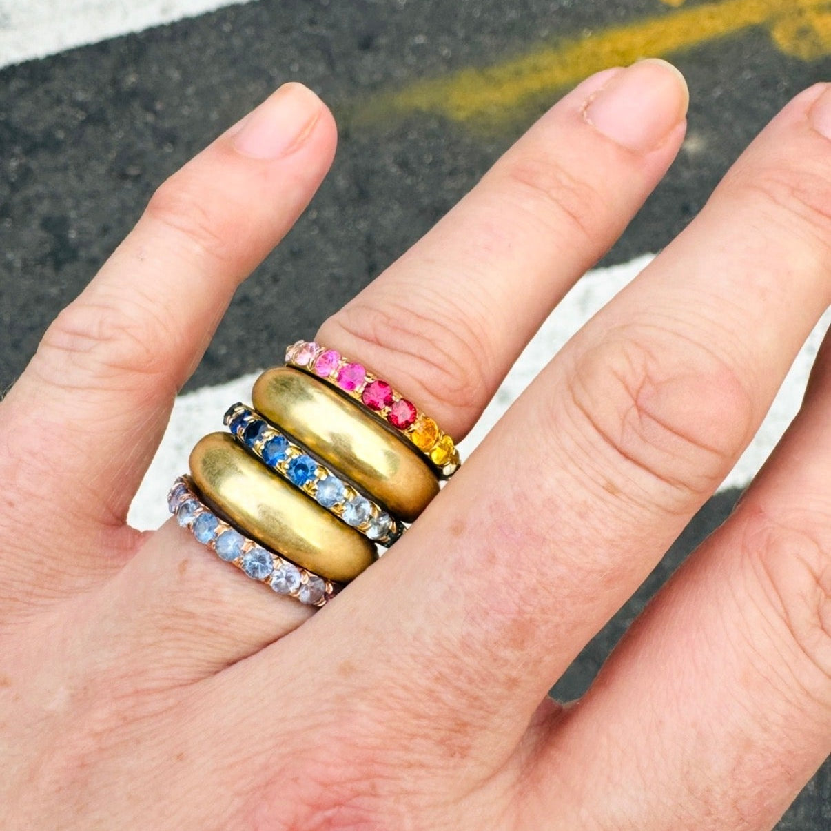 A close-up of a hand wearing a stack of five rings, including two thick gold bands with smaller accent rings featuring pink, blue, and clear stones, set against an asphalt background with painted white lines.