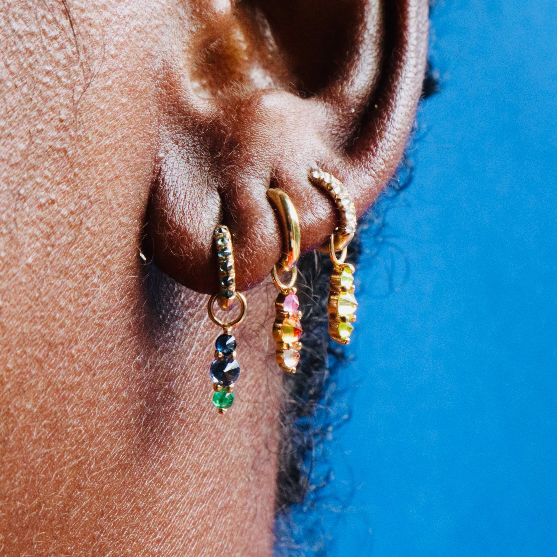 Three Mini Barbed Diamond Chunky huggie hoop earrings with colorful gemstone charms on models ear against blue background