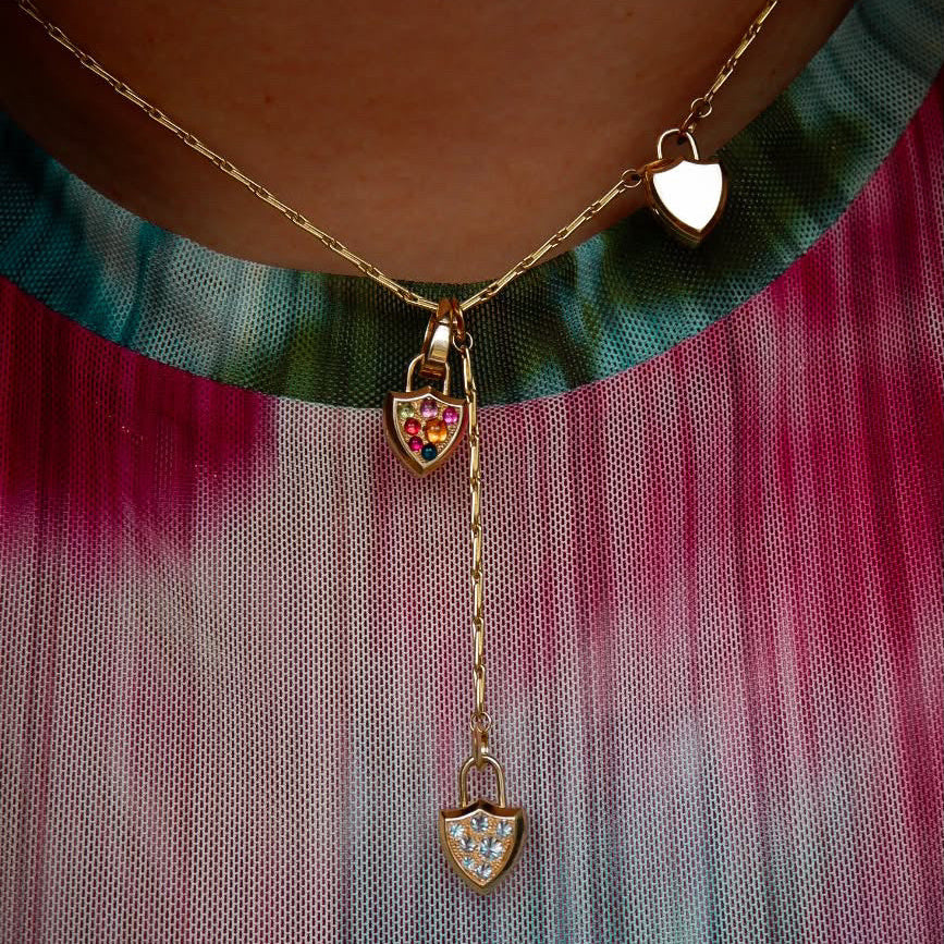 A close-up of a gold necklace worn on a person's neck, featuring a Y-shaped chain design with three small shield shaped locket pendants, one solid gold, one gold with diamond pavé and one gold with rainbow cabochon pavé, over a teal & pink tie-dye mesh shirt.