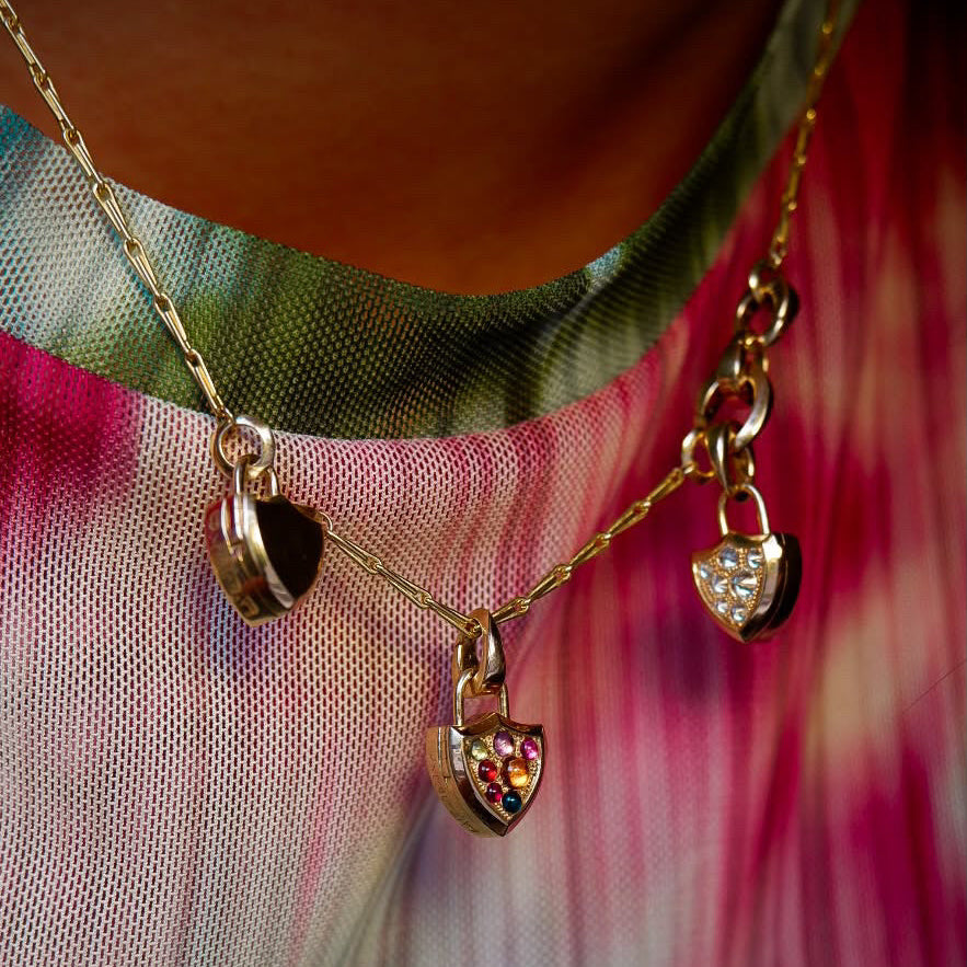 A close-up of a gold necklace worn on a person's neck, featuring a Y-shaped chain design with three small shield shaped locket pendants, one solid gold, one gold with diamond pavé and one gold with rainbow cabochon pavé, over a teal & pink tie-dye mesh shirt.