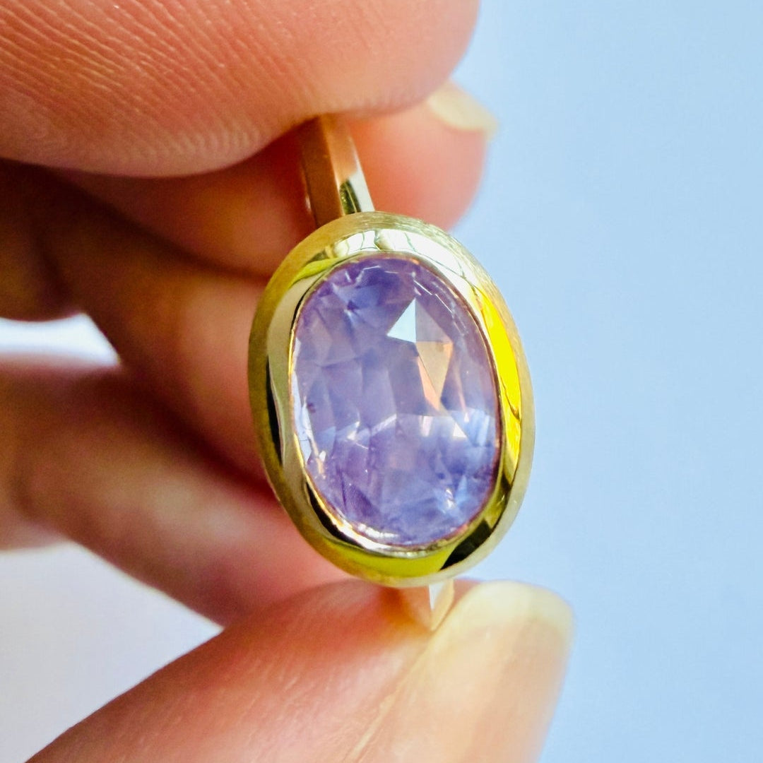 Gold ring with a purple sapphire gemstone held between fingers against a light blue background