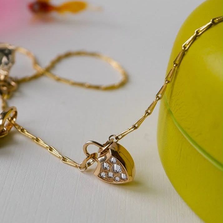 A close-up of a gold pendant necklace , featuring a barley corn chain design with a shield shaped locket pendant in gold with spiked diamond pavé against a cream background surrounded by some blurred but colorful props