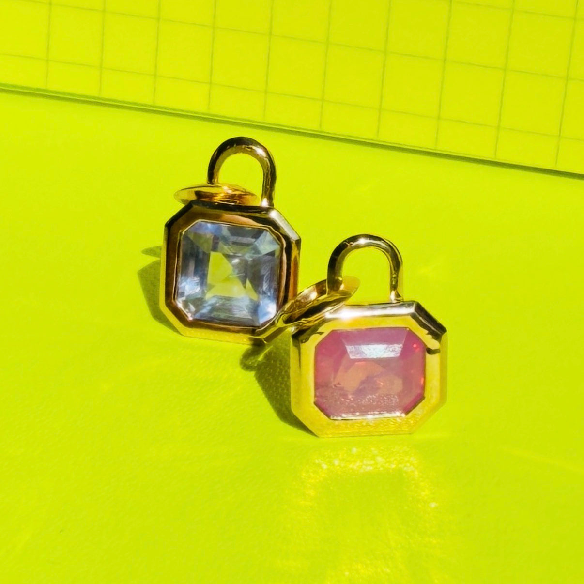 One pink spinel and  one lavender sapphire bezel set gemstone charms, displayed on a textured neon yellow surface by Tamsin Rasor.