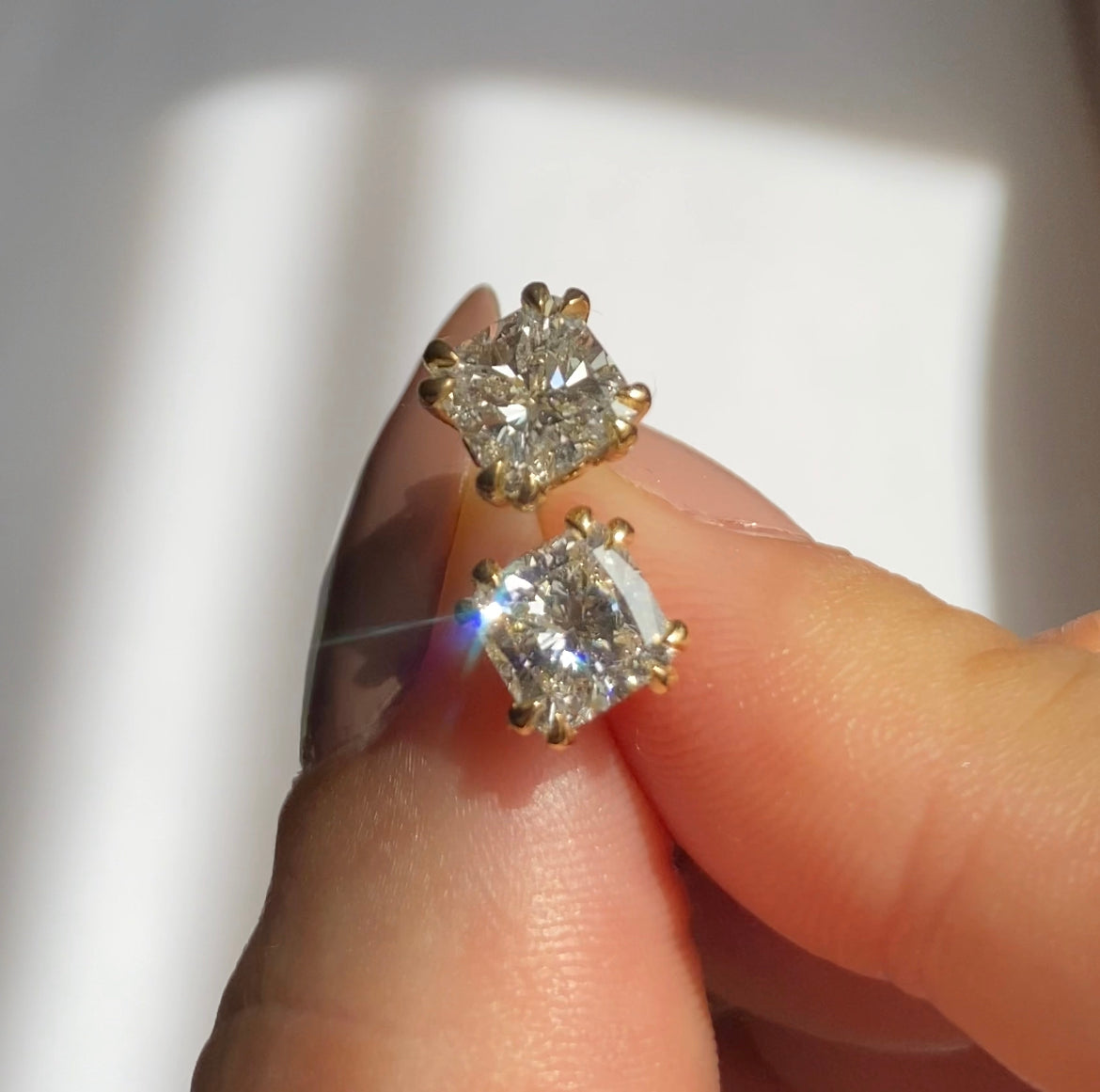A pair of cushion cut white diamond earrings set in 18k yellow gold held between two fingers on white background