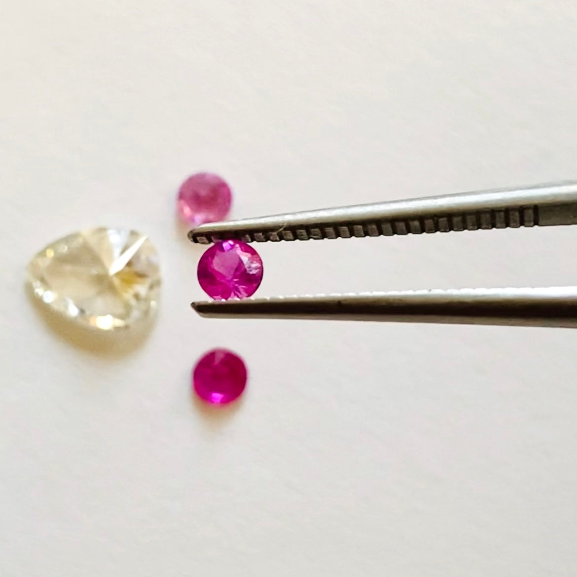 Round Pink Sapphire held in tweezers above pear shaped white diamond and two round fuchsia pink sapphire gem stones shown on white background
