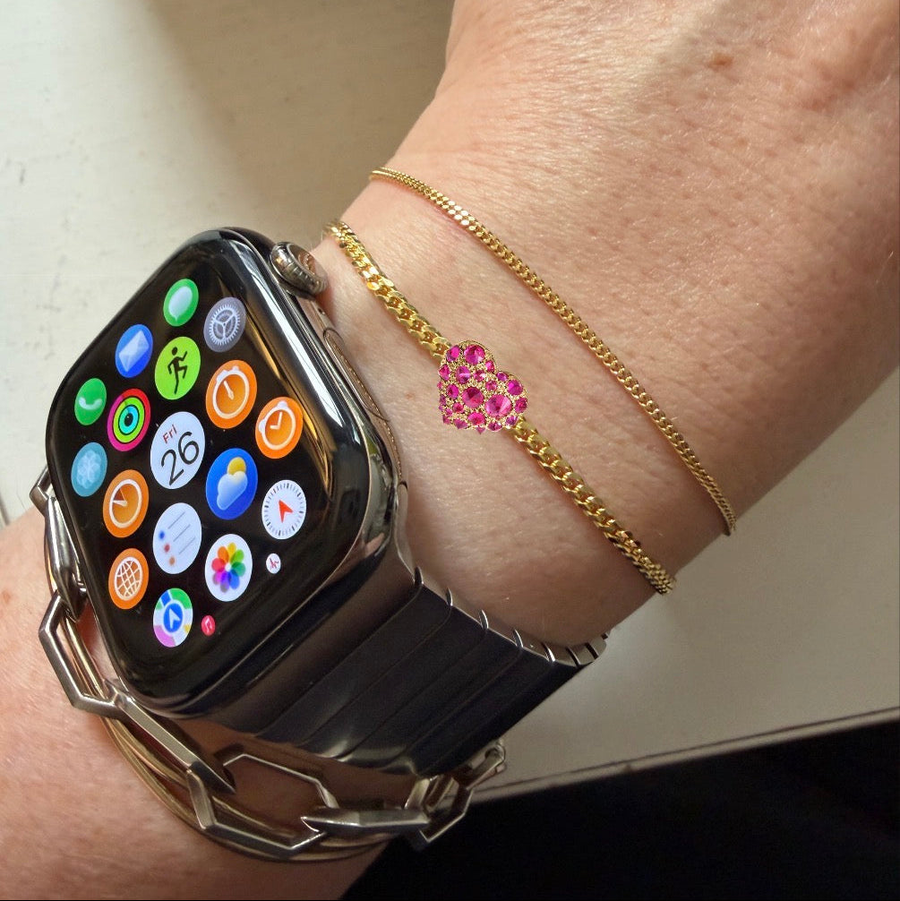 Gold bracelet with a heart-shaped ruby charm by Tamsin Rasor shown on models wrist stacked with gold chain bracelets and a smart watch