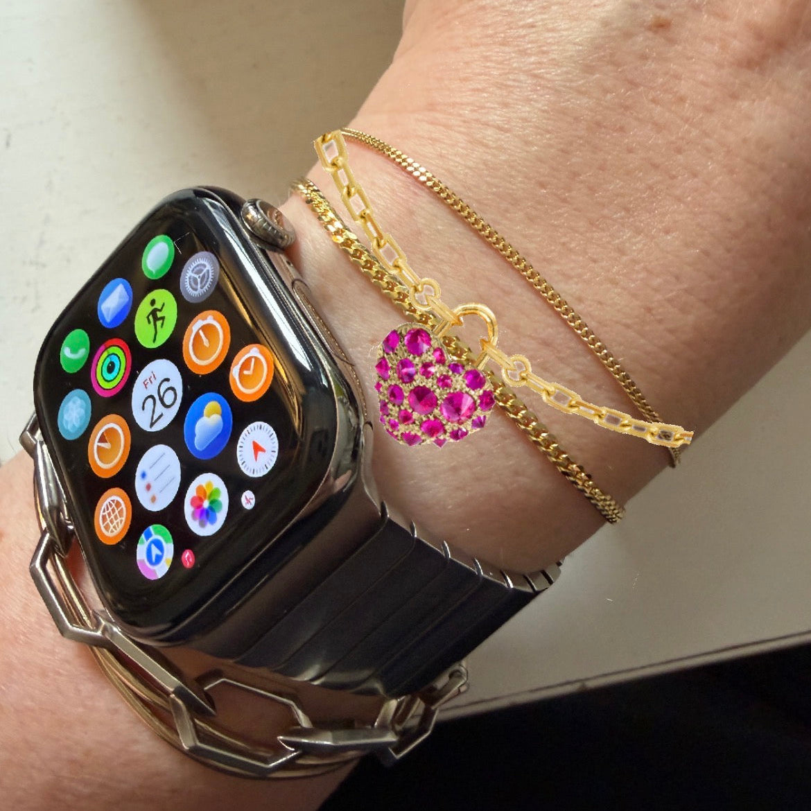 Gold bracelet with a heart-shaped ruby charm by Tamsin Rasor shown on models wrist stacked with gold chain bracelets and a smart watch