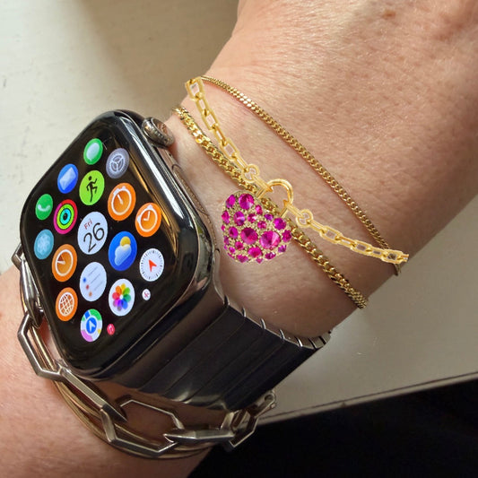 Gold bracelet with a heart-shaped ruby charm by Tamsin Rasor shown on models wrist stacked with gold chain bracelets and a smart watch