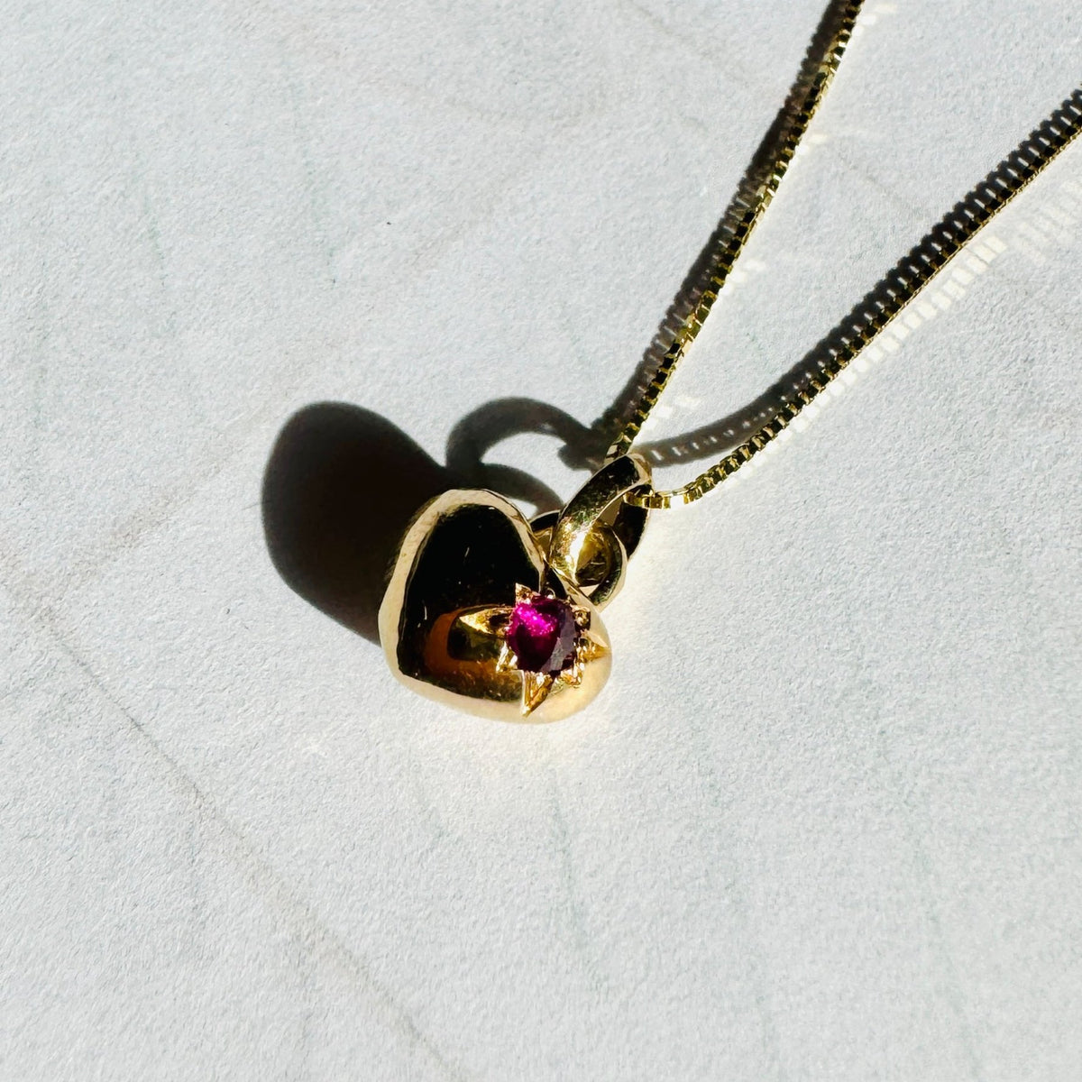 Gold heart-shaped pendant on a chain,  with a single star set spiked ruby gemstone on the right hand side, against a white background