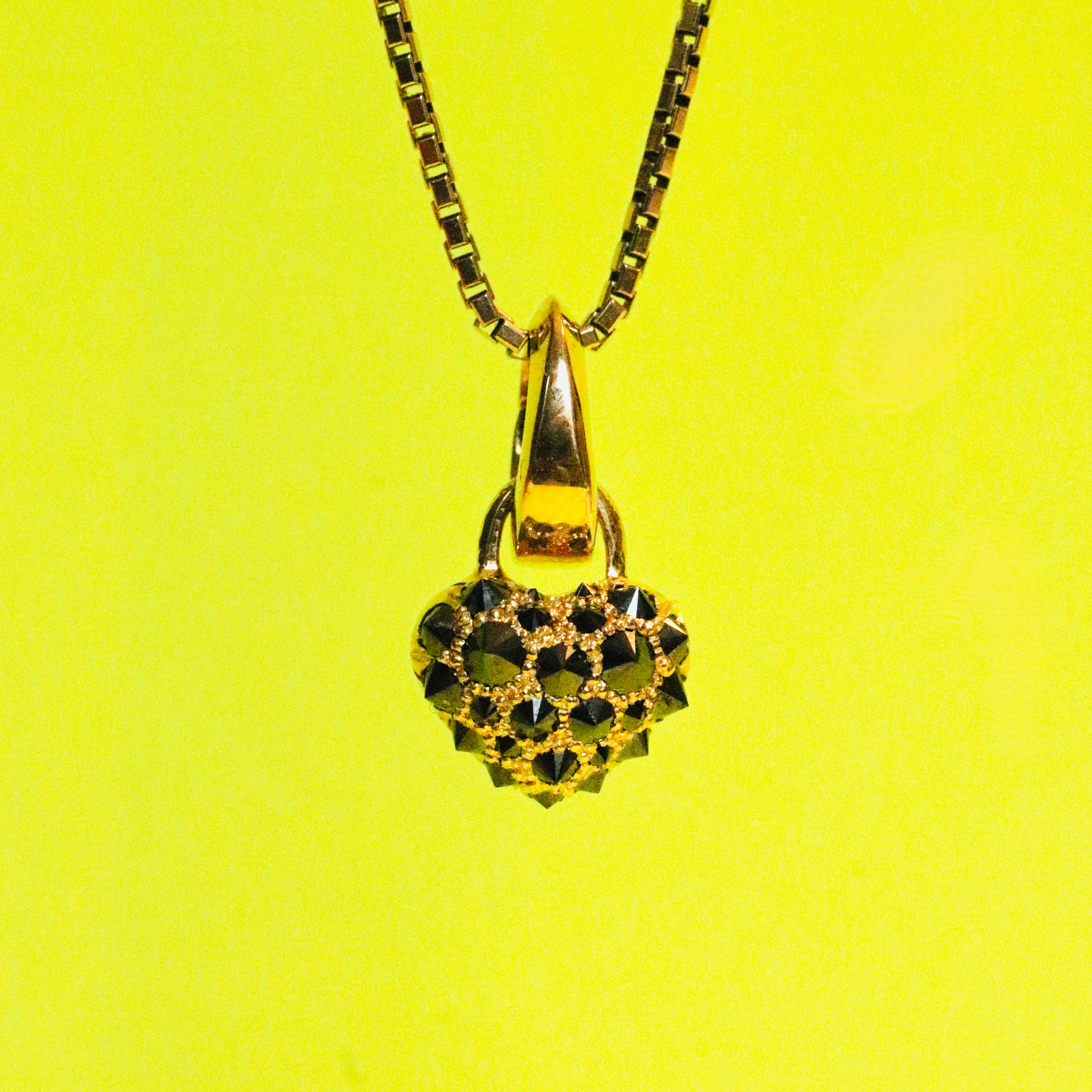 Heart charm set with upside down spiked black diamonds by Tamsin Rasor in 18K gold , hangin from a gold box chain, in front of a neon yellow background 