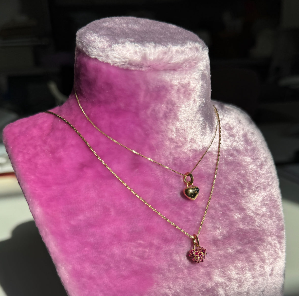 Two gold heart-shaped pendants on chains. One with a single star set spiked black diaomond and one covered in spiky ruby pavé shown on a pink velvet neckform