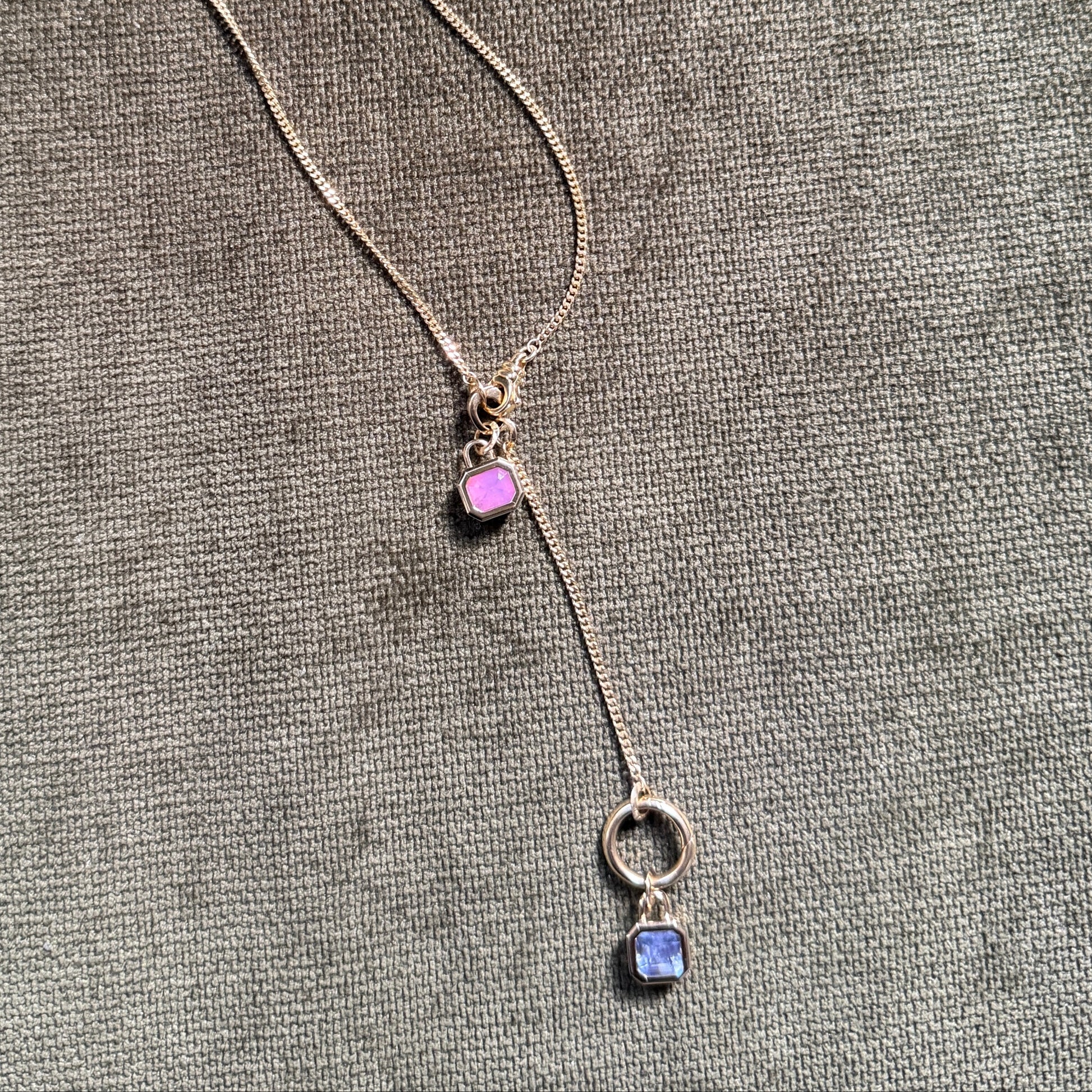 bezel set gemstone pendants on gold chain with charm clips. One pin Spinel and one lavender sapphire. Against olive green velvet background