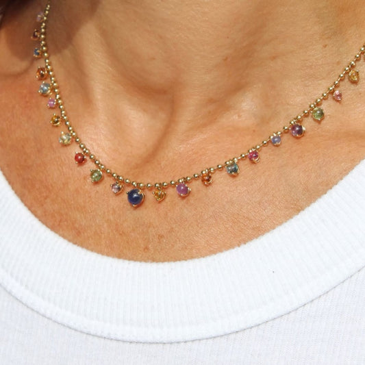 A close up of a gold bead chain necklace featuring a fringe of multi color sapphire cabochon gem drops, set in gold prong settings,By Tamsin Rasor, shown around models neck. model is wearing a white t-shirt. 