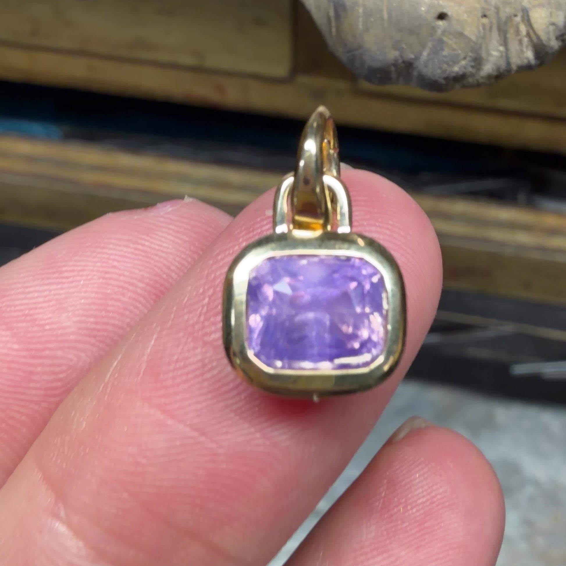 video of a bezel set lavender sapphire and 18K gold pendant in modes hand, shown in front of a jewelers bench