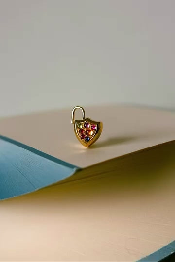 stop motion film of small gold shield shaped locket with padlock top bail opening and closing . Locket is sat on top of a beige and blue book and is covered in rainbow colored cabochon gemstones