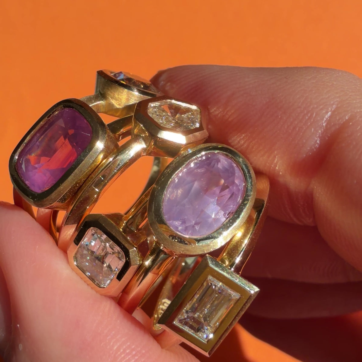 Gold, diamond and pink sapphire gemstone rings by Tamsin Rasor held between model's fingers against orange background