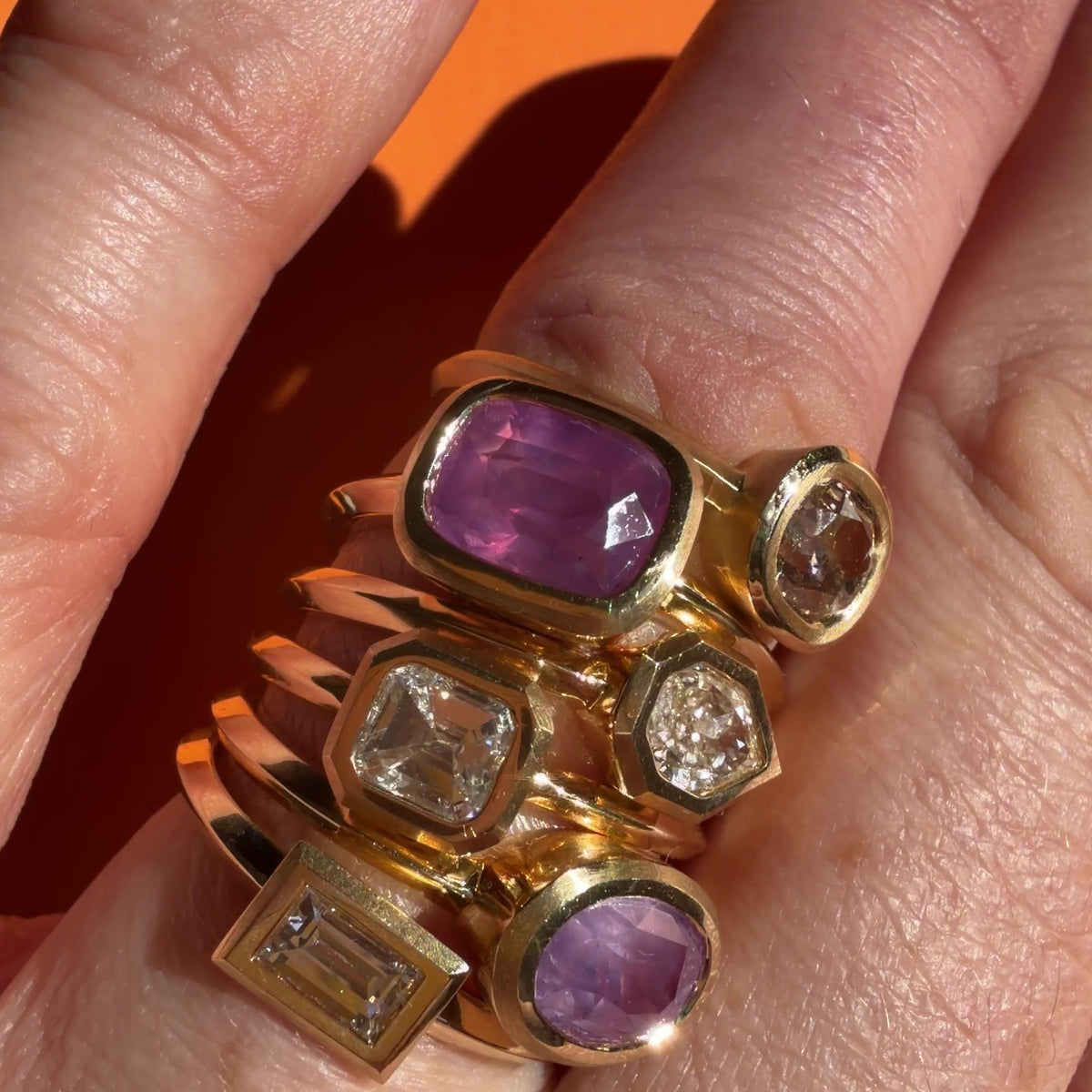 Six sapphire and diamond rings by Tamsin Rasor, stacked on models finger against orange background
