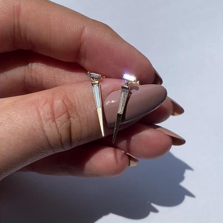 A pair of yellow gold baguette cut diamond drop earrings with a push-back closure on in models hand, against white background, by Tamsin Rasor.